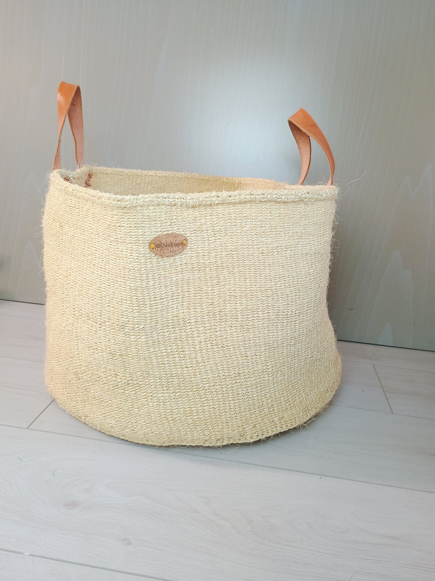Beige woven basket with leather handles on a light wooden floor.